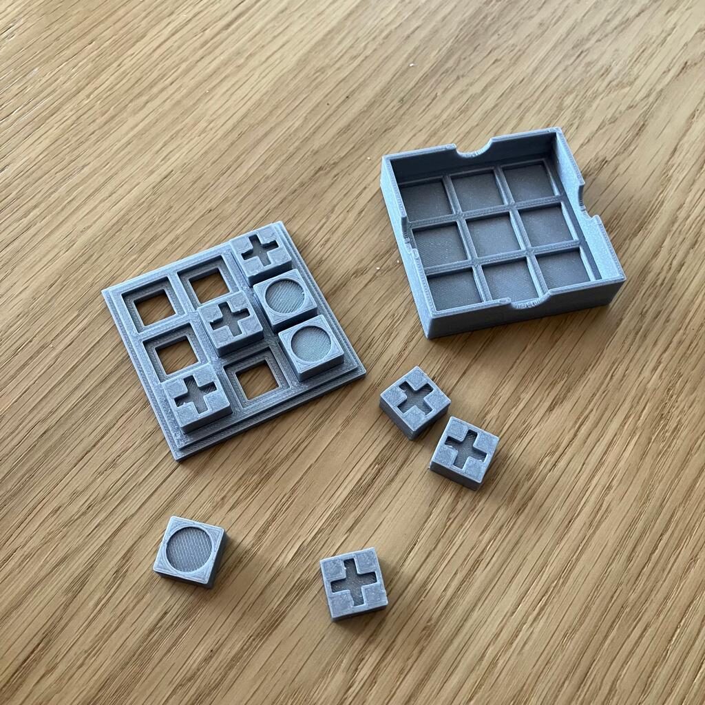 Kółko i krzyżyk, figury i plansza Kółko i krzyżyk - stół, gra leży w dwóch częściach, obok rozsypane cztery kostki: 3 z krzyżykiem, jedna z kółkiem. Na planszy ułożona skośna linia z trzech krzyżyków