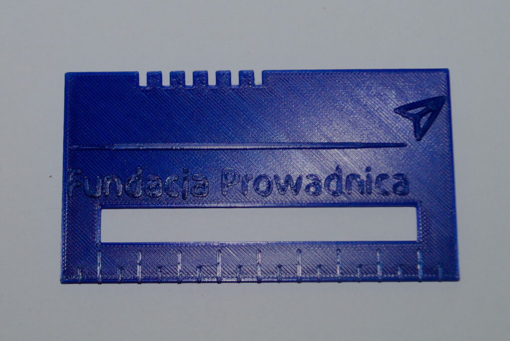 Ramka prowadząca, z logo, ABS niebieski Niebieska ramka z miejscem na podpis. Na krawędziach znajdują się wypukłe logo Fundacji Prowadnica oraz linijka. Na dole wklęsła banknotówka.