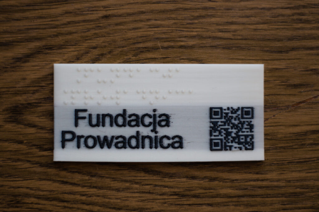 PLA, biała tabliczka z czarnym napisem Fundacja Prowadnica, białym napisem brajlowskim i kodem QR Na górze tabliczki znajduje się brajlowski napis "Fundacja Prowadnica", podzielony na dwa rzędy. Pod nim analogiczny napis czarnymi, czarnodrukowymi literami. Z prawej strony umieszczony jest kod QR.