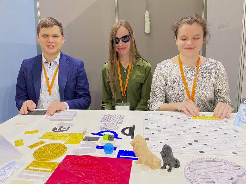 TargiOpole2 Drugi dzień Targów Dostępności. Stoisko widoczne jest z perspektywy gościa. Na stole leżą różne rekwizyty, przy nim siedzą Dawid, Monika oraz Julita.