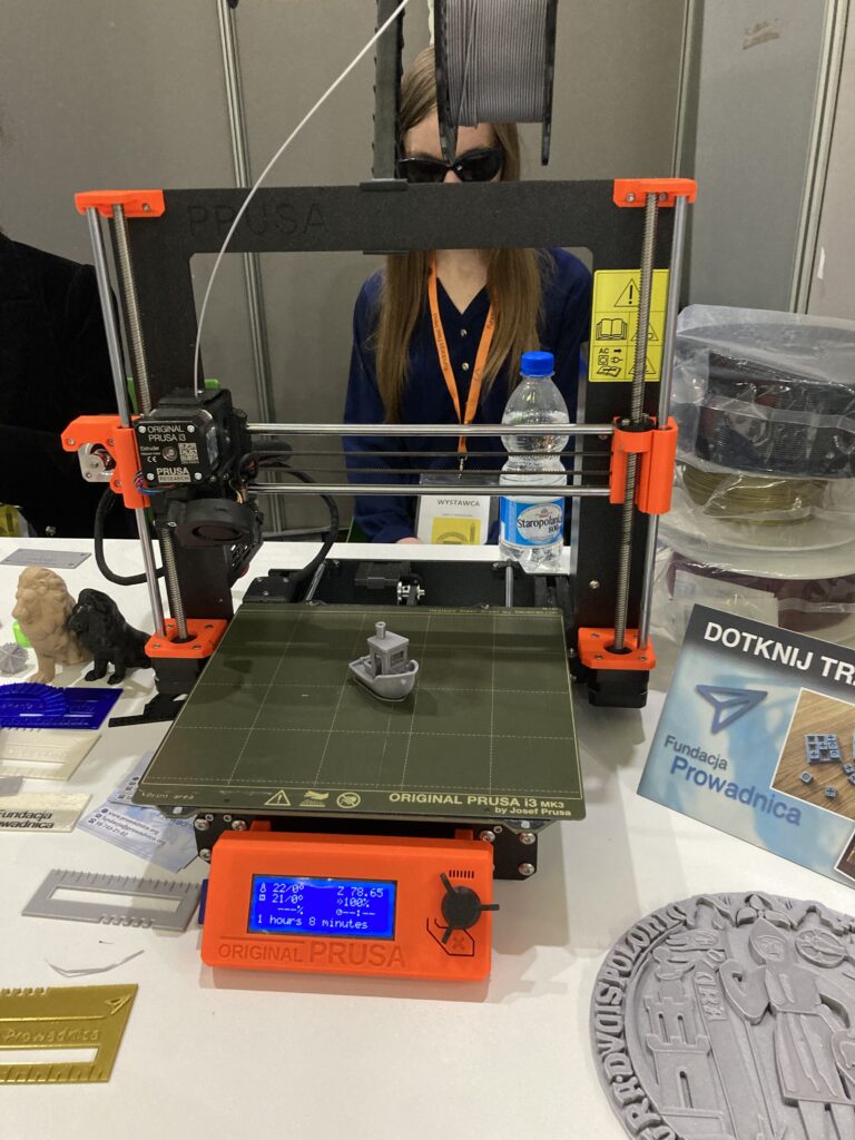 TargiOpole6 Targi Dostępności. Widać drukarkę 3d wykonującą tzw. 3d Benchy, czyli łódkę, ze srebrnego filamentu. Za drukarką siedzi Monika.