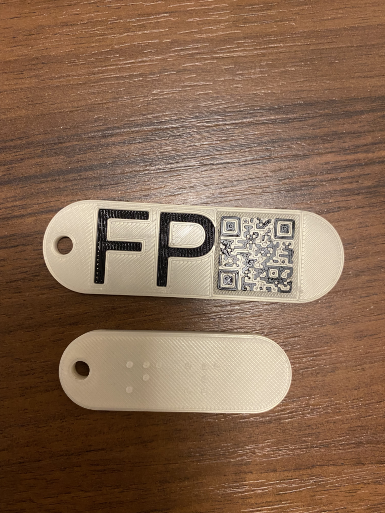 Breloczki: czarnodruk, Brajl i kod QR Dwa białe breloczki: na górnej powierzchni jednego widnieją brajlowskie litery FP, na drugiego czarny napis FP i kod QR.