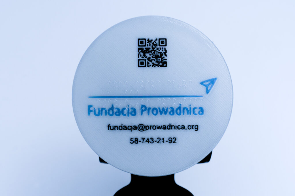 Tabliczka z Brajlem i poddrukiem Koło z brajlowskim napisem Fundacja Prowadnica, w poddruku kod QR do strony Fundacji, logo i dane kontaktowe
