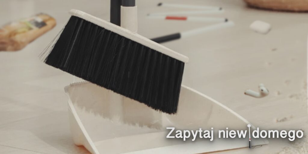 Głównymi obiektami na grafice są zmiotka i szufelka. Stoją skierowane w stronę zmiecionej kubki śmieci na podłodze. W tle widać nogę od stołu i duży wazon. Na podłodze wala się też na wpół złożona biała laska, otwarta kanapka z Żabki i etui Airpods pro. Gdzieś dalej od etui leży jedna słuchawka.