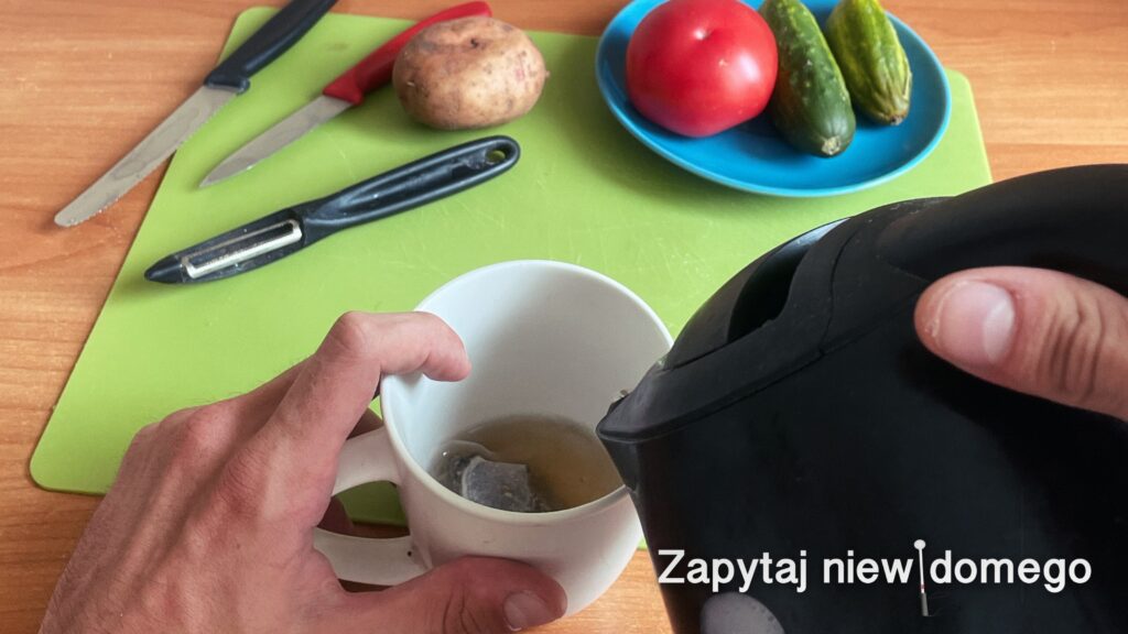 Widok stołu z zieloną podkładką. Na pierwszym planie dłonie, nasz grafik przyznał, że jego własne, ;) wlewają wrzątek do kubka: prawa trzyma czajnik, palec wskazujący lewej oparty jest o wewnętrzną stronę kubka. Dalej, po lewej, leżą ostrzami do widza dwa noże: jeden domasła, drugi ostrzejszy i obieraczka. Obok obieraczki nieobrany ziemniak. Po prawej stoi talerz z pomidorem i dwoma ogórkami.
