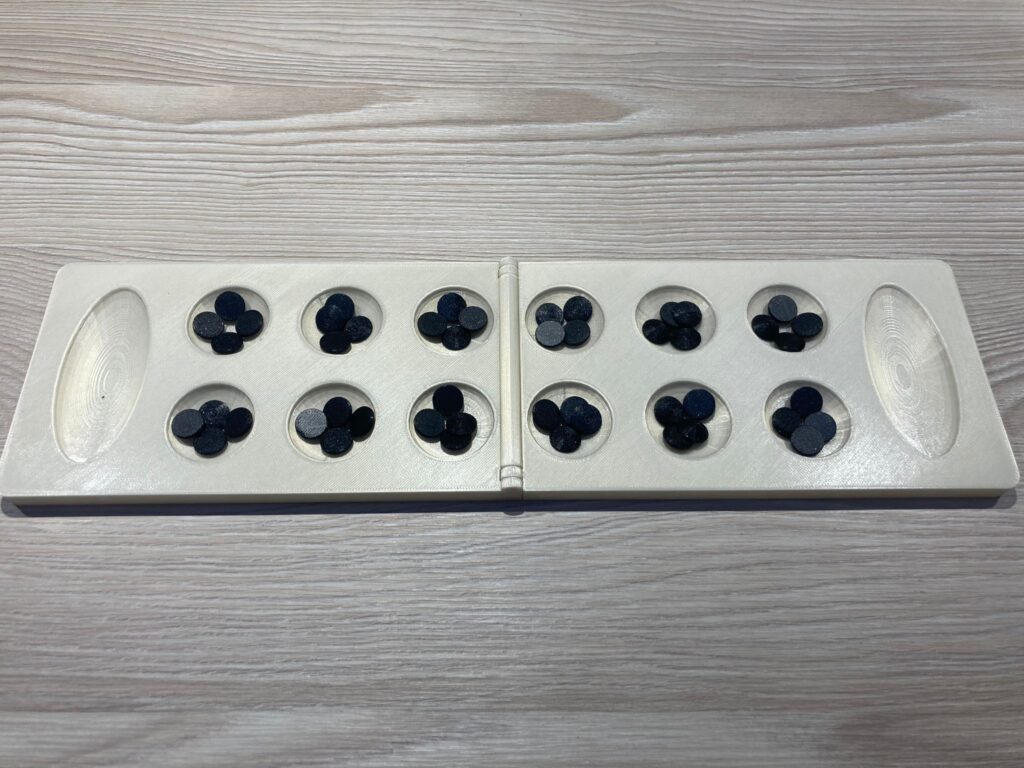 mankala z daleka, biała plansza i czarne kamienie Biała plansza mankali składa się z dwunastu (po sześć na przeciwko siebie) półkolistych otworów oraz, po bokach, dwóch podłużnych. W połowie szerokości znajduje się zawias umożliwiający jej złożenie.