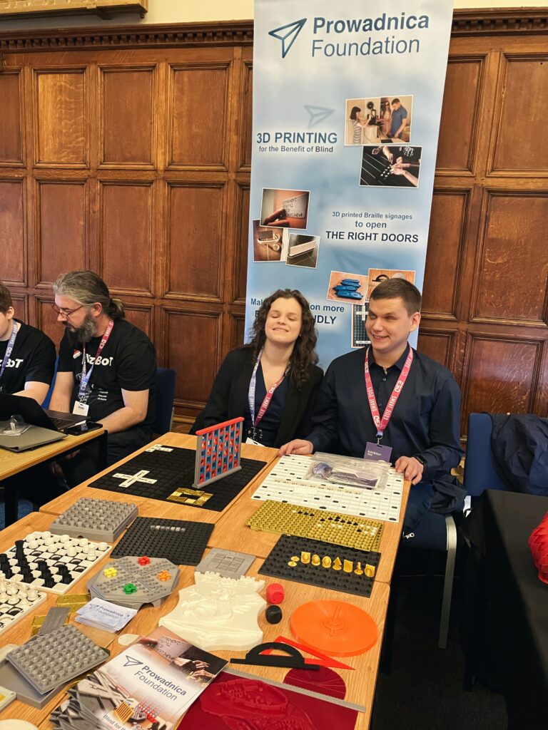 widok stanowiska fundacji lekko pod kątem, Uśmiechnięta Julita schyla głowę ku Dawidowi; widać baner fundacji i stół z wydrukami