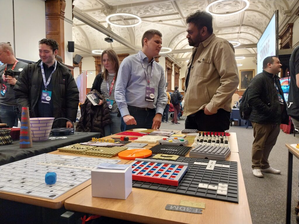 Na pierwszym planie stół z wydrukami. Za nim Dawid pokazuje wydruki zwiedzającemu mężczyźnie