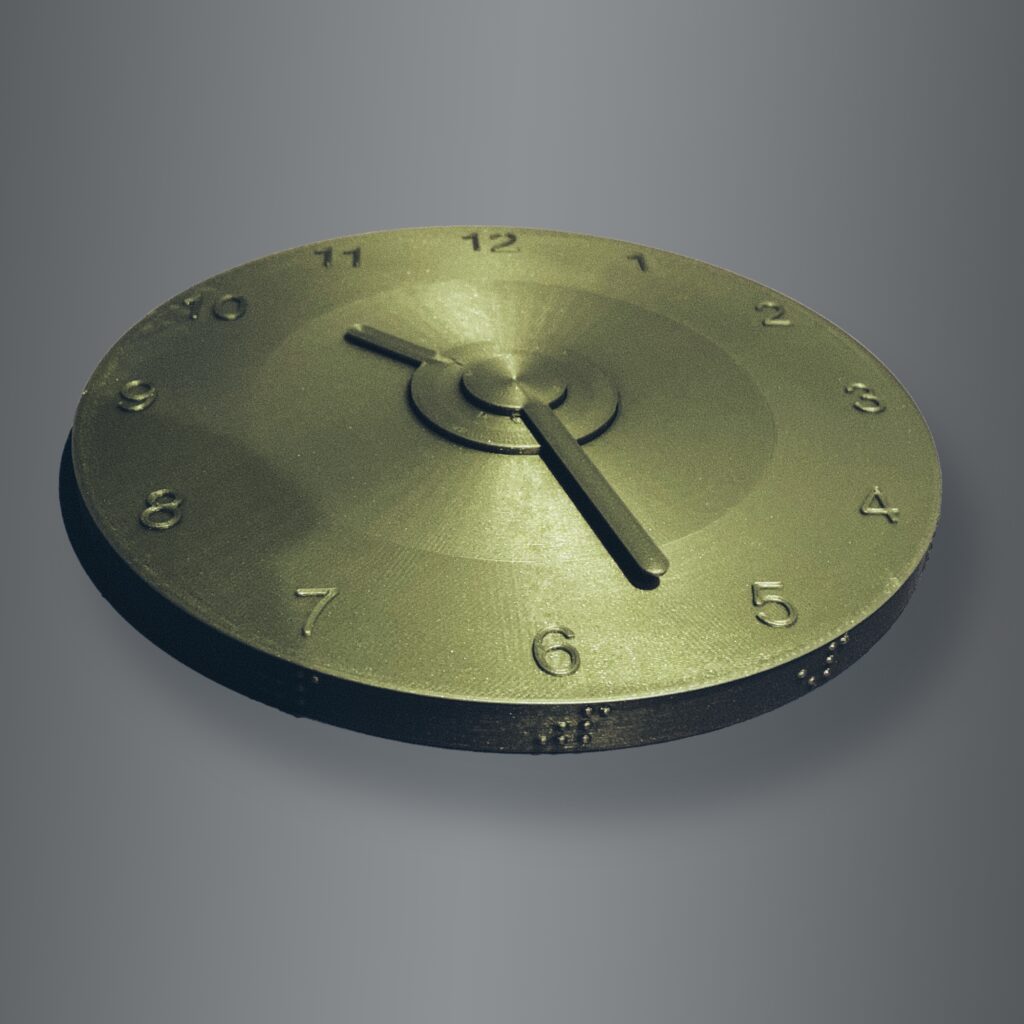 Zegar pod kątem Złota tarcza zegara widziana pod kątem, na niej dwie, obracane wskazówki. Na górze tarczy znajdują się czarnodrukowe oznaczenia godzin, na krawędziach brajlowskie.