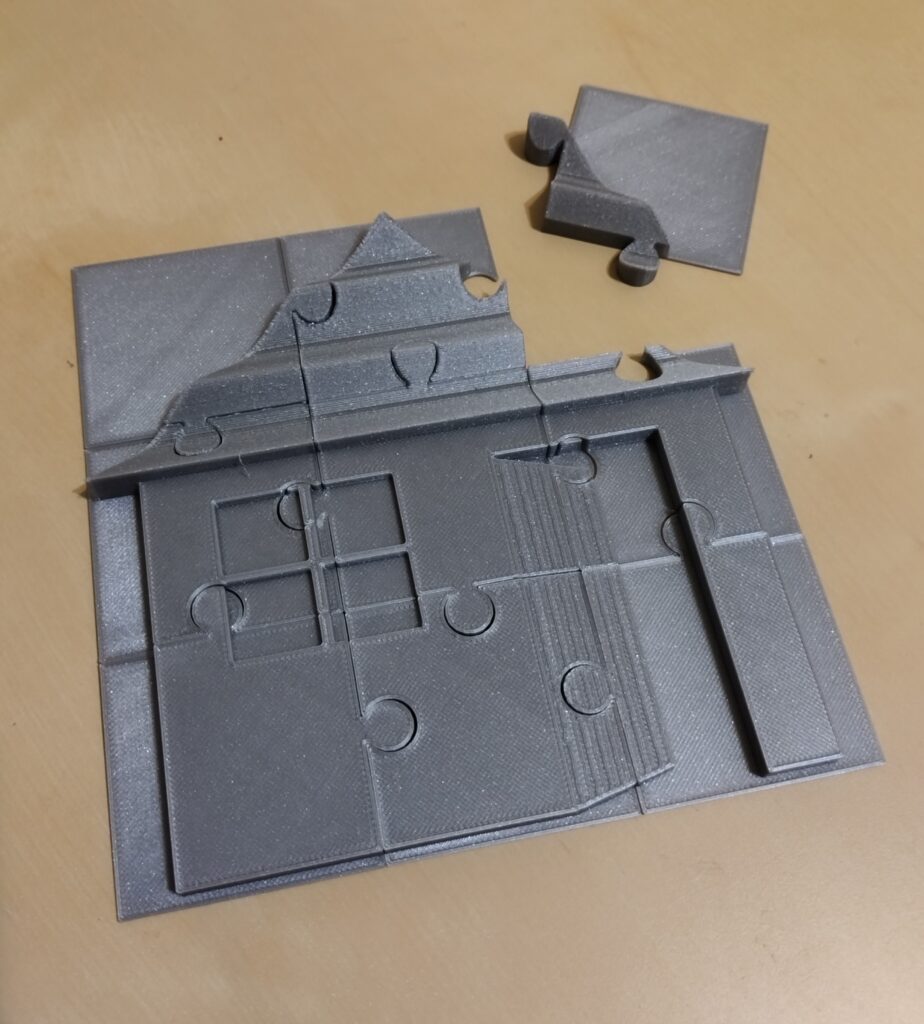 Puzzle Domek Domek składany z puzzli (3 na 3 elementy). Z lewej strony znajdują się cztery okna na planie kwadratu, z prawej uchylone drzwi. Domek przykrywa falisty dach.